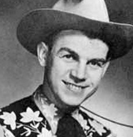 A smiling young man wears a wide-brimmed cowboy hat and a patterned shirt with floral designs, posing for a black-and-white portrait.