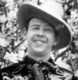 A smiling person wearing a wide-brimmed cowboy hat and a neckerchief, dressed in western-style clothing, with trees in the background. The image is in black and white.