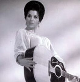 A woman with dark hair styled up is holding a large acoustic guitar and wearing a light-colored blouse. The photo is in black and white, and she is looking slightly to the side.
