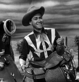 A smiling person in a cowboy hat and western shirt holds a saddle, standing outdoors with a cloudy sky and ranch equipment visible in the background.