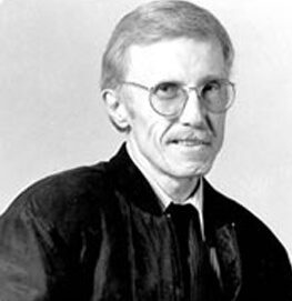 A black-and-white portrait of an older man with short, light hair wearing round glasses, a mustache, a dark jacket, and a collared shirt. He is facing slightly to the side and looking at the camera.