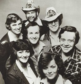 A black and white photo of seven adults, five men and two women, smiling and posing closely together. Two men are wearing cowboy hats. The group appears cheerful and friendly.