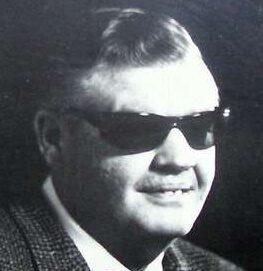 A man with short hair wearing dark sunglasses and a checkered jacket, smiling slightly in a black and white portrait.