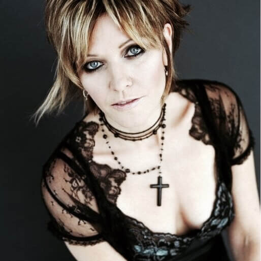 A woman with short, layered blonde hair wears a black lace top and a black cross necklace. She gazes upward at the camera with a neutral expression against a dark background.