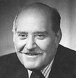 Black-and-white portrait of an older man with a bald head and mustache, smiling, wearing a suit and tie.