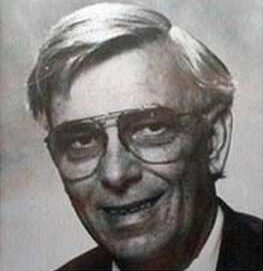 Black and white portrait of an older man with light hair, wearing large eyeglasses and a suit jacket, smiling slightly while looking at the camera.