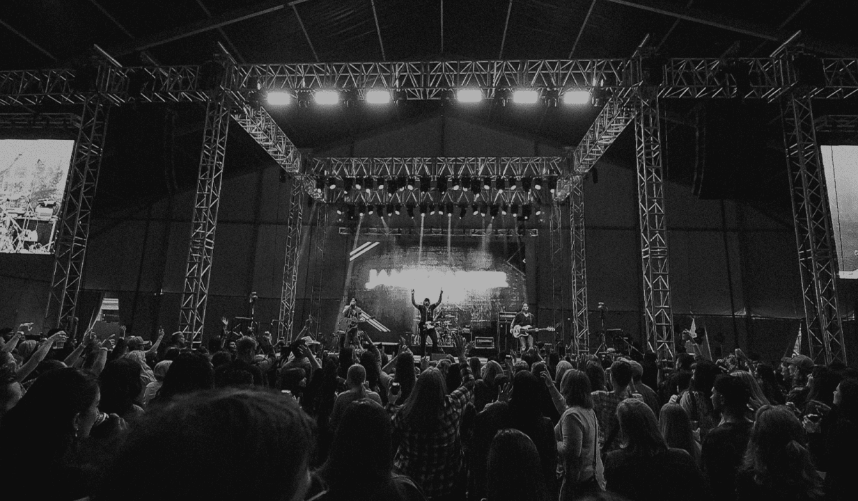 A band performs on a truss-framed indoor festival stage, with a large energetic crowd cheering and raising their hands.
