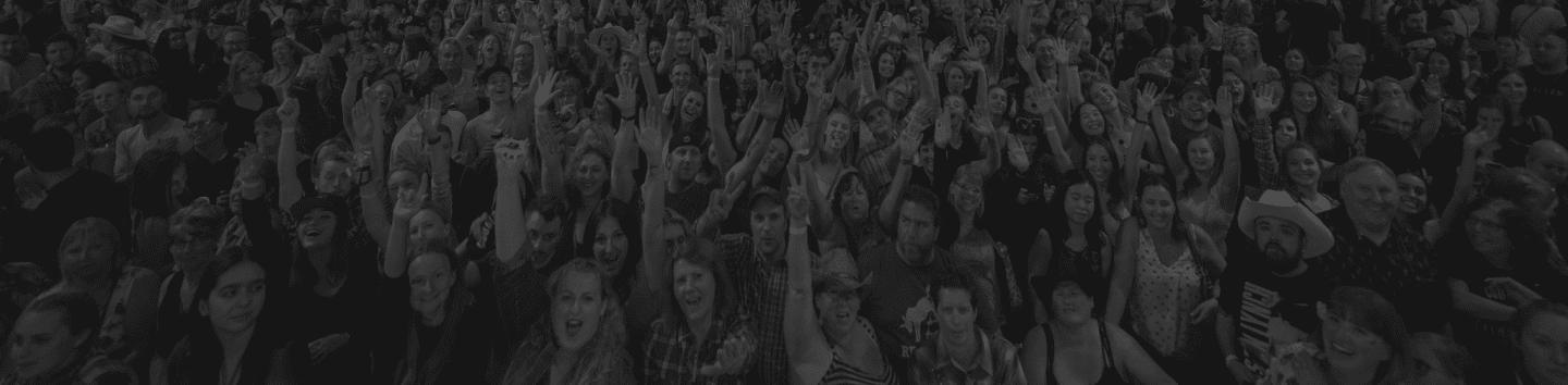 A large, energetic crowd of concert-goers cheering with hands raised, photographed from above in black and white