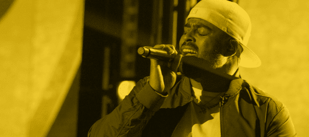 A male performer sings passionately into a microphone wearing a white beanie and jacket, bathed in warm golden stage lighting
