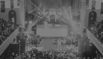 A performer on a lit stage in an ornate indoor venue, surrounded by a packed crowd on both the main floor and upper balconies
