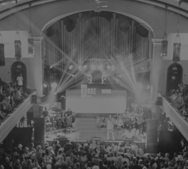 A performer on a lit stage in an ornate indoor venue, surrounded by a packed crowd on both the main floor and upper balconies